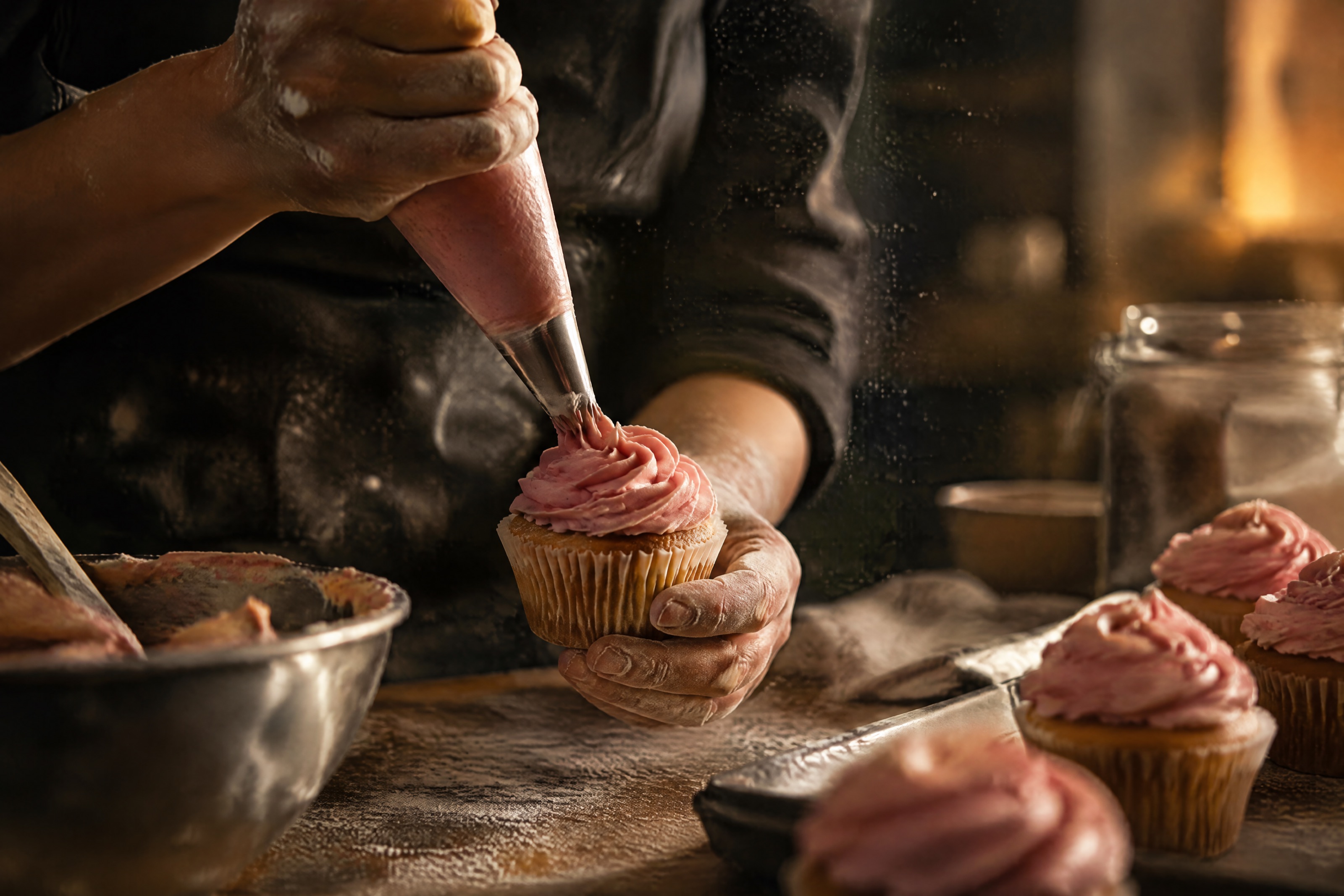 A baker's hands piping pink buttercream onto a cupcake in a warm, flour-dusted kitchen